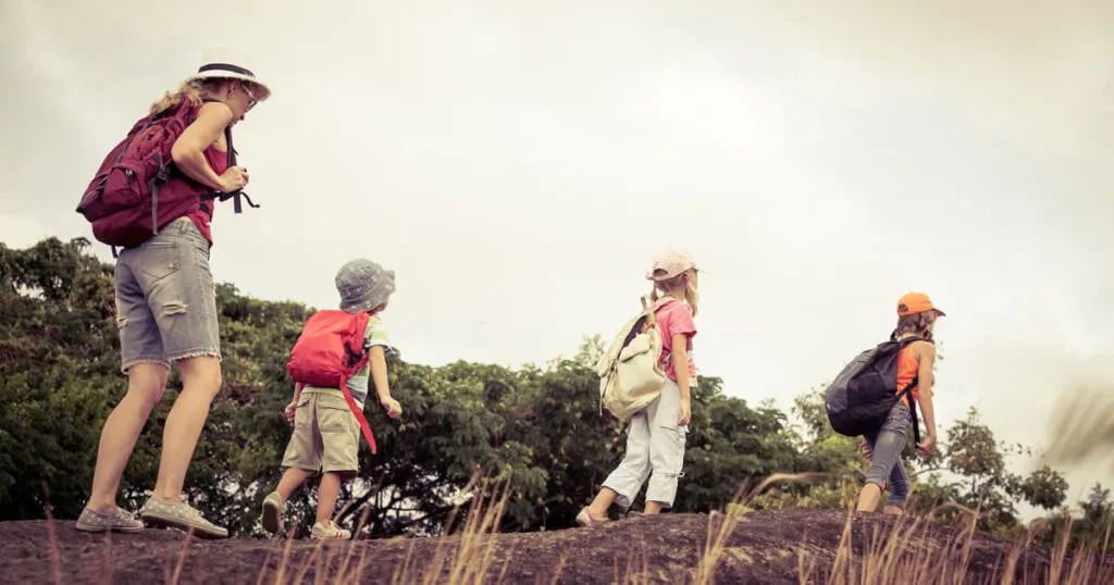 A woman is going for a hike with her 3 children. They are all wearing summer clothes, sunhats, running shoes, and a small daypack. They are walking on a log, one behind the other. The mother is in the back, following the lead of her children.